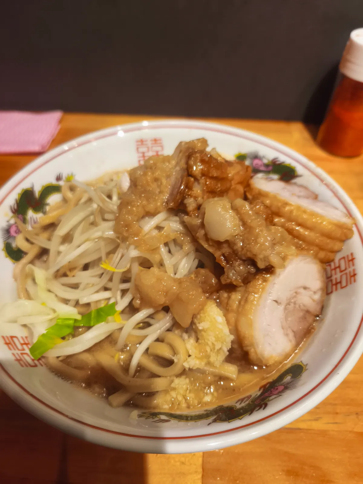 ブタゴリラのブタタマラーメン。龍が描かれた丼に、もやし中心の野菜、極太のうねった麺、背脂の塊、おろし生姜の山、そして大きな豚が2枚のっている。スープは醤油の色が濃い茶色