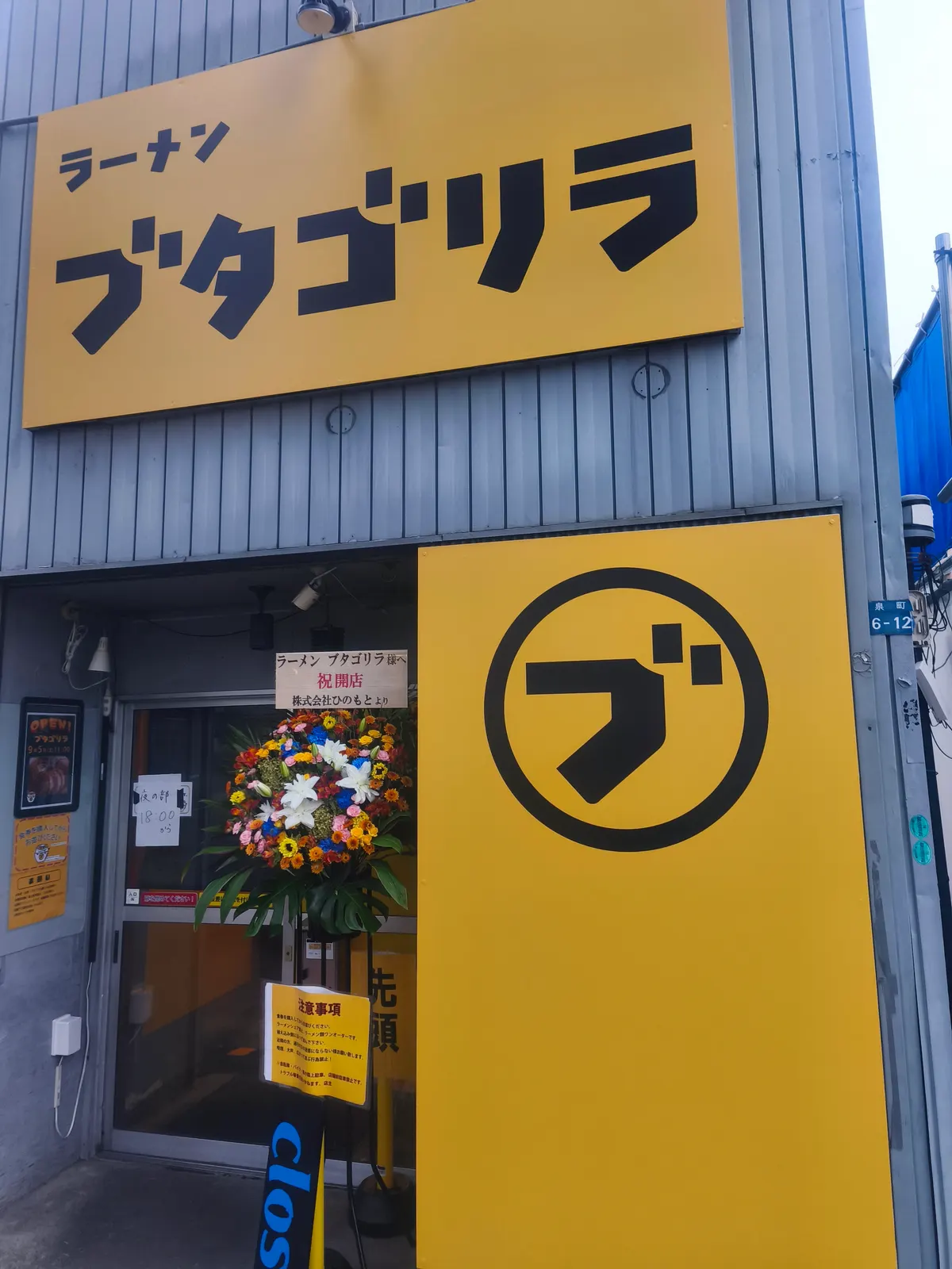 ブタゴリラの外観。黄色地に黒文字の「ラーメン ブタゴリラ」の看板と、丸に「ブ」のロゴが入った黄色いシャッター。入口には開店祝いの花スタンドが置かれている
