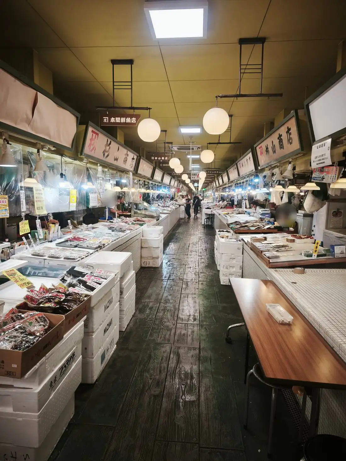 はこだて自由市場の場内通路。両側に鮮魚店が並び、丸い照明が下がっている。床は濡れている