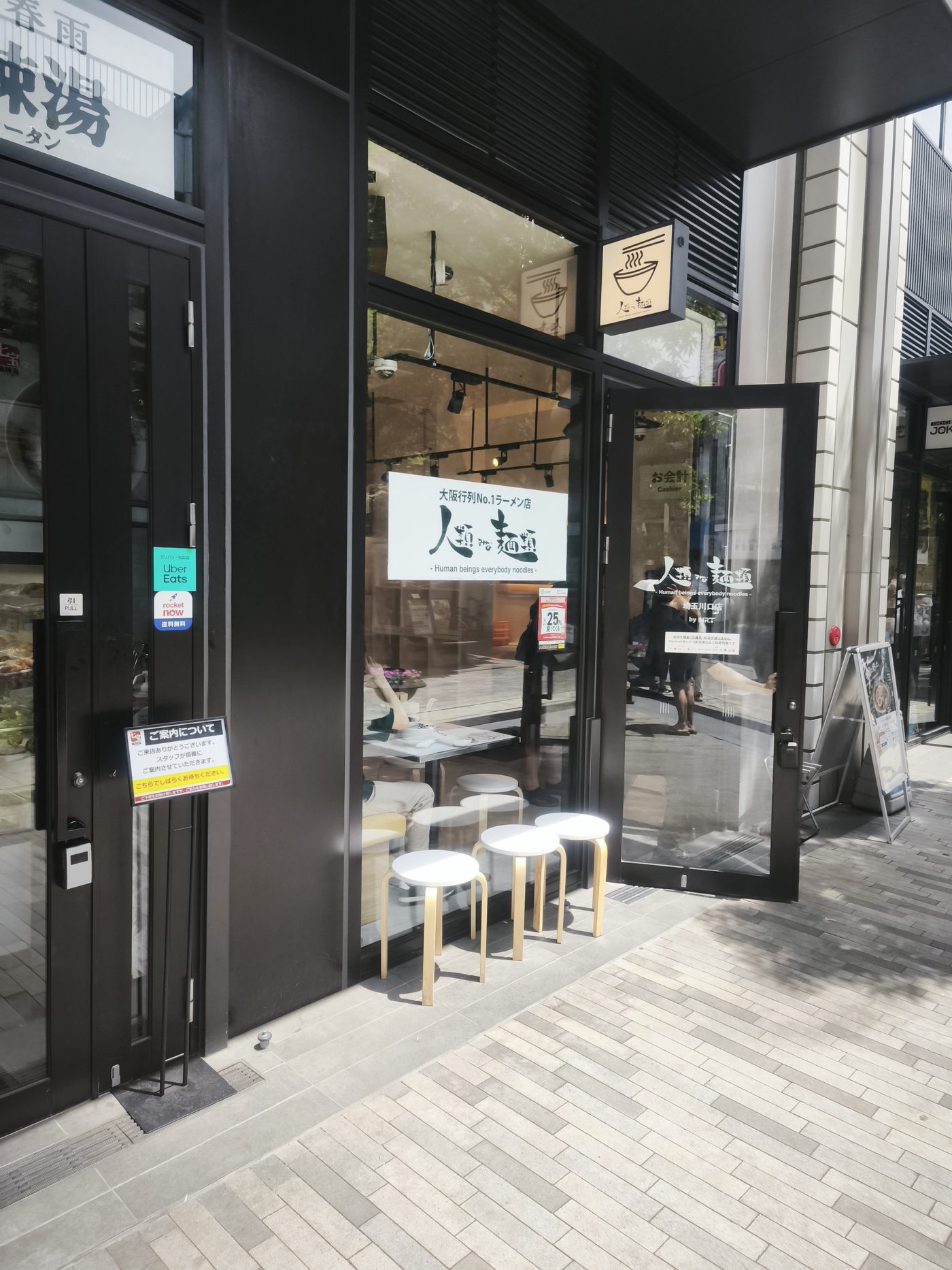 人類みな麺類 埼玉川口店の外観。ガラス張りの店構えと行列用の丸イスが並ぶ