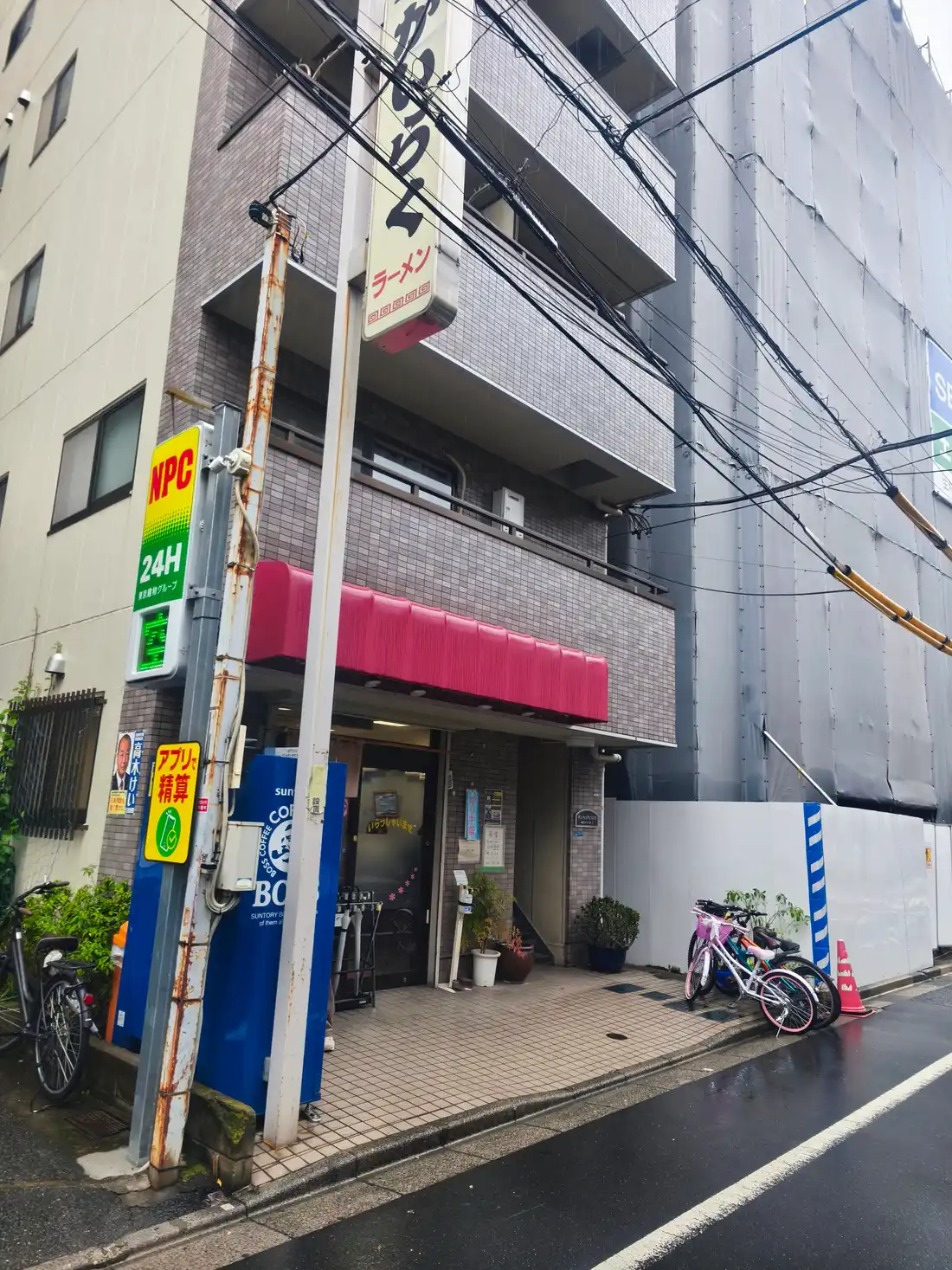 かいらくの外観。雑居ビルの1階、赤い庇(ひさし)に「ラーメン」の文字。雨の日の路地で、店先には自転車が停まっている