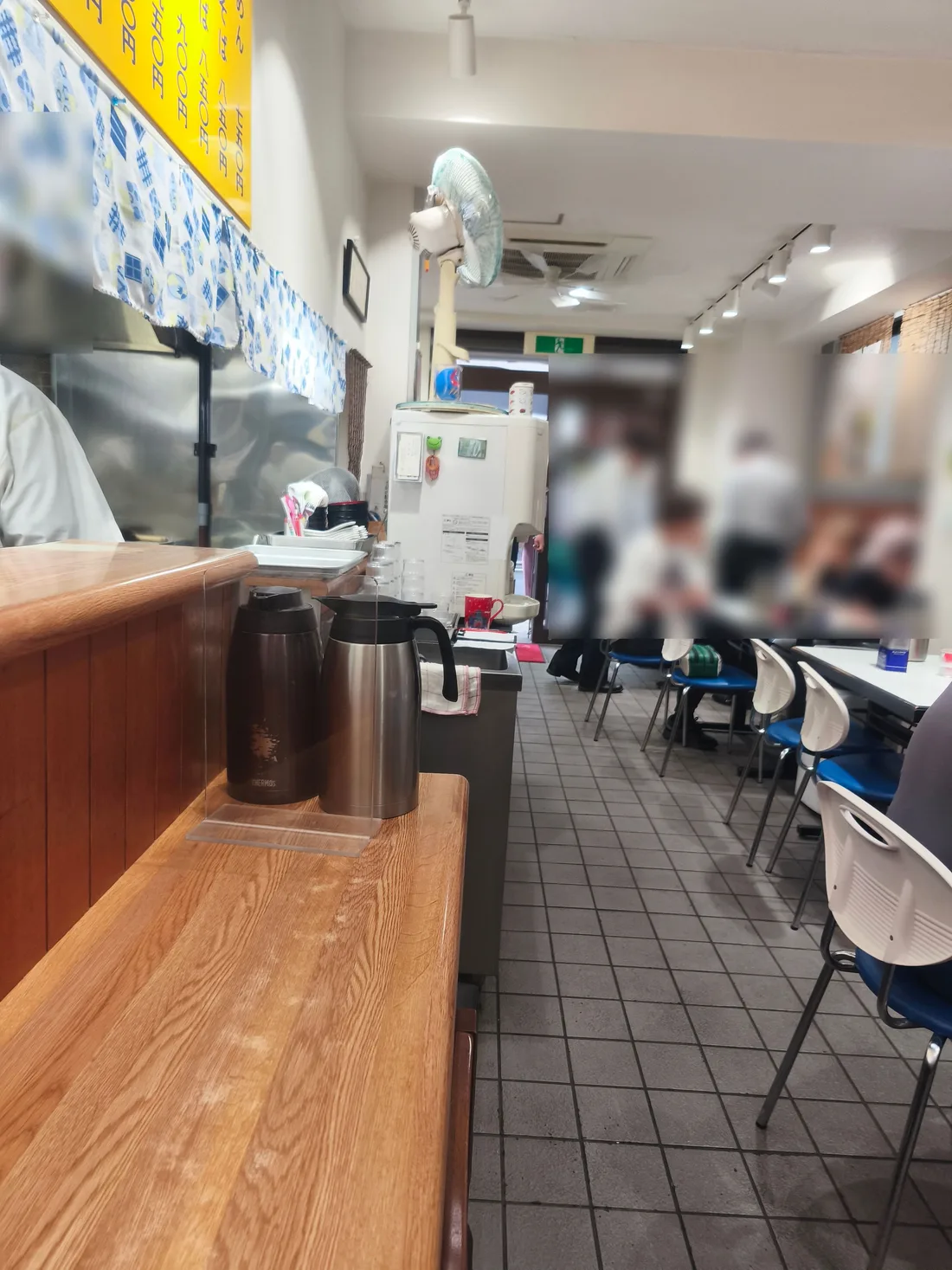 かいらくの店内。細長い造りで、右側にテーブル席、左側が厨房。手前のカウンターにはステンレスのポットが並ぶ(他のお客さんが写っているためぼかしています)